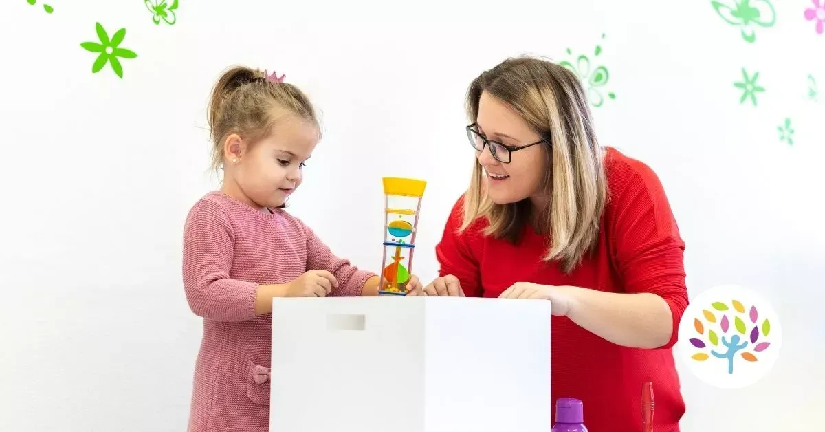 A child and an adult interact while playing with a small, colorful toy on a white table against a floral-patterned wall.
