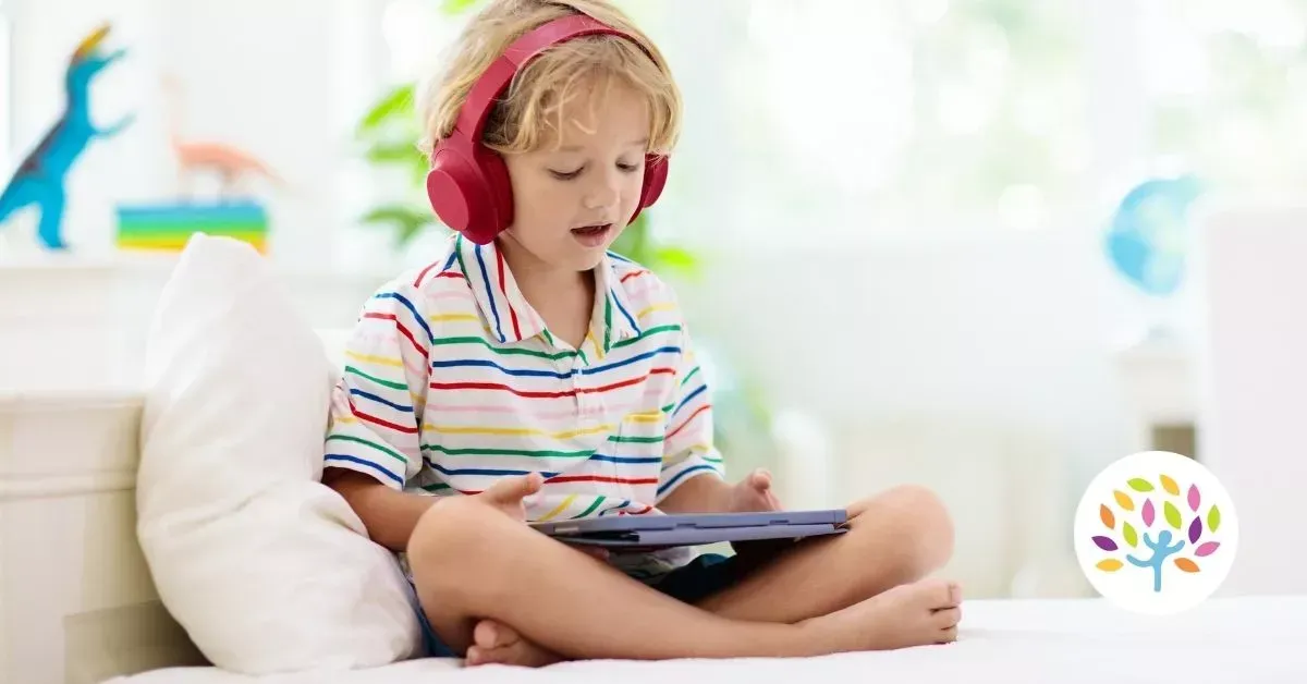 A young person wearing red headphones sits on a bed, looking at a tablet in a bright, colorful room.