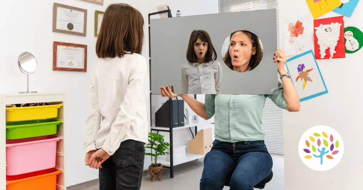 A therapist holds a mirror and a facial expression card for a student in a room with colorful bins and wall decorations.