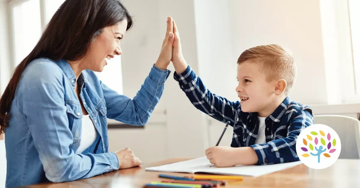 A smiling adult and child high-five while sitting at a desk with an open book and colorful pencils.