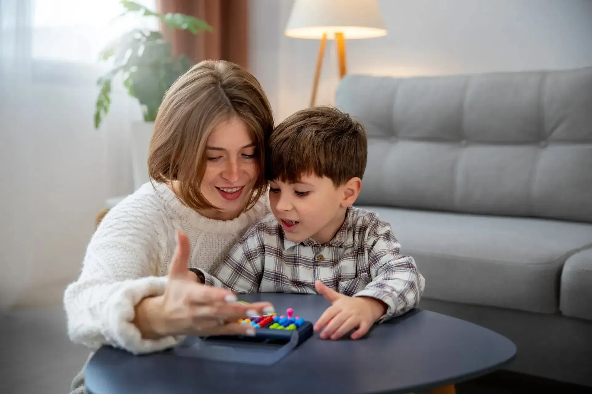 A person and child sit together at a table, smiling while playing a game with small, colorful pieces in a bright room.