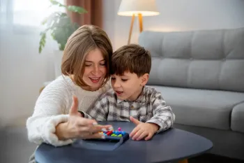 A person and child sit together at a table, smiling while playing a game with small, colorful pieces in a bright room.