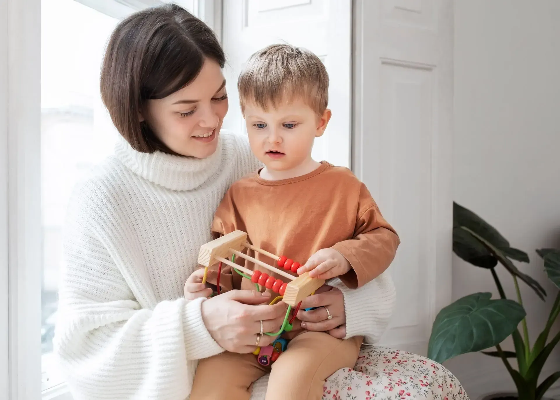 A person in a white sweater sitting by a window, holding a young child who is playing with a small wooden bead toy.