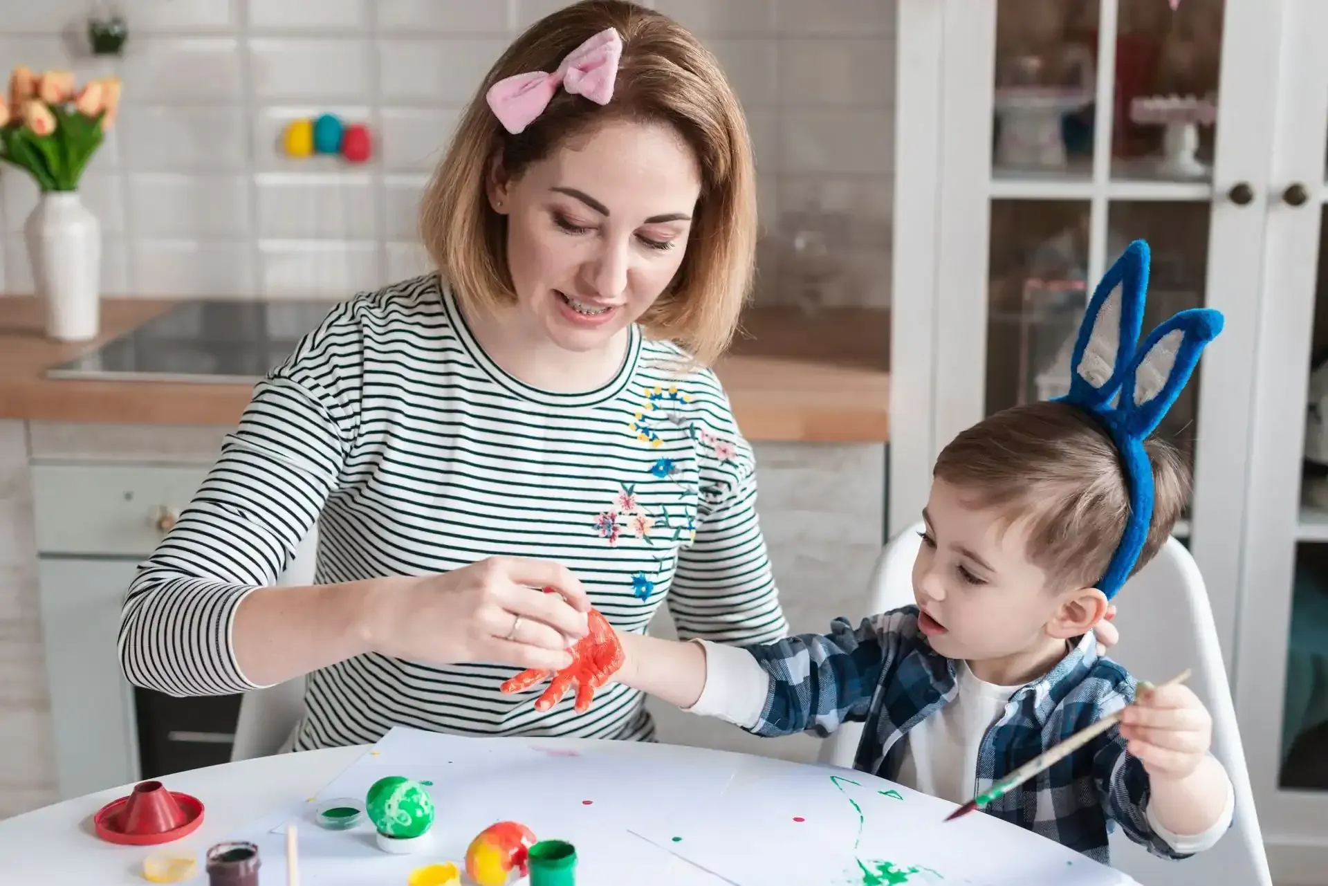 A person with a pink bow painting a child's hand with red paint at a table filled with art supplies in a kitchen.
