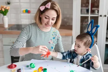 A person with a pink bow painting a child's hand with red paint at a table filled with art supplies in a kitchen.