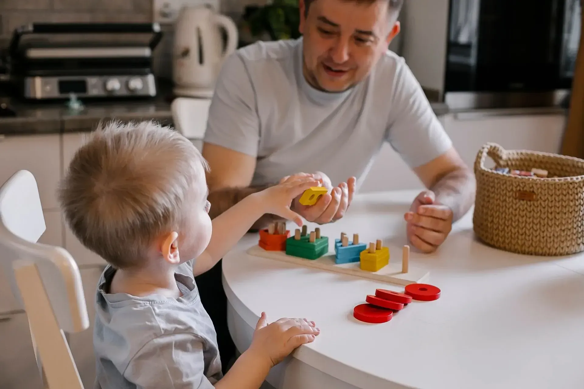 A parent and child sitting at a kitchen table playing with a colorful wooden shape sorting toy.