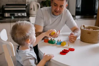 A parent and child sitting at a kitchen table playing with a colorful wooden shape sorting toy.