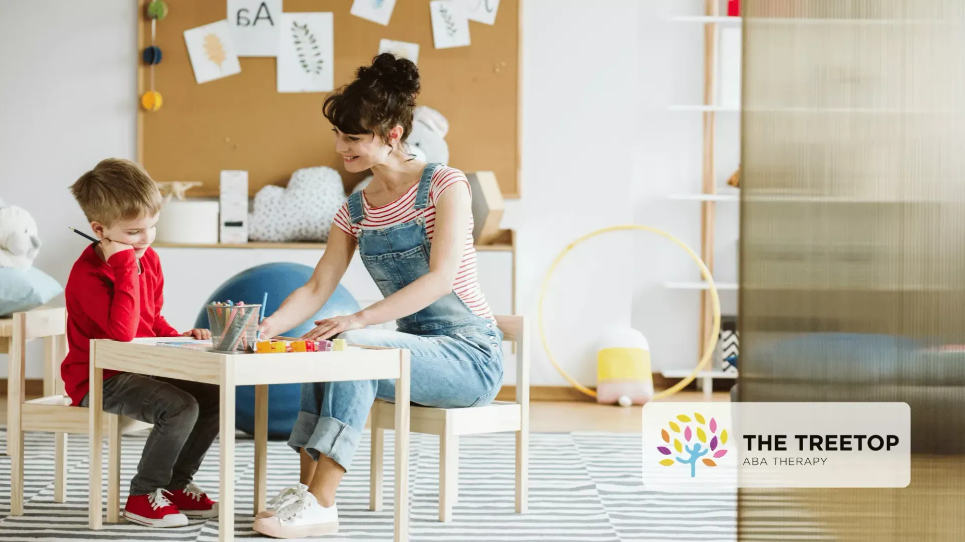 A teacher sits at a small table with a student in a bright, organized classroom, engaging in a hands-on learning activity.