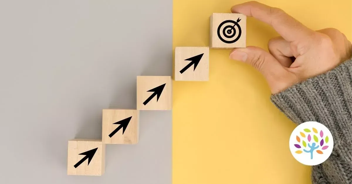 A hand places a wooden block with a target icon onto a stair-step formation of blocks with upward-pointing arrows.