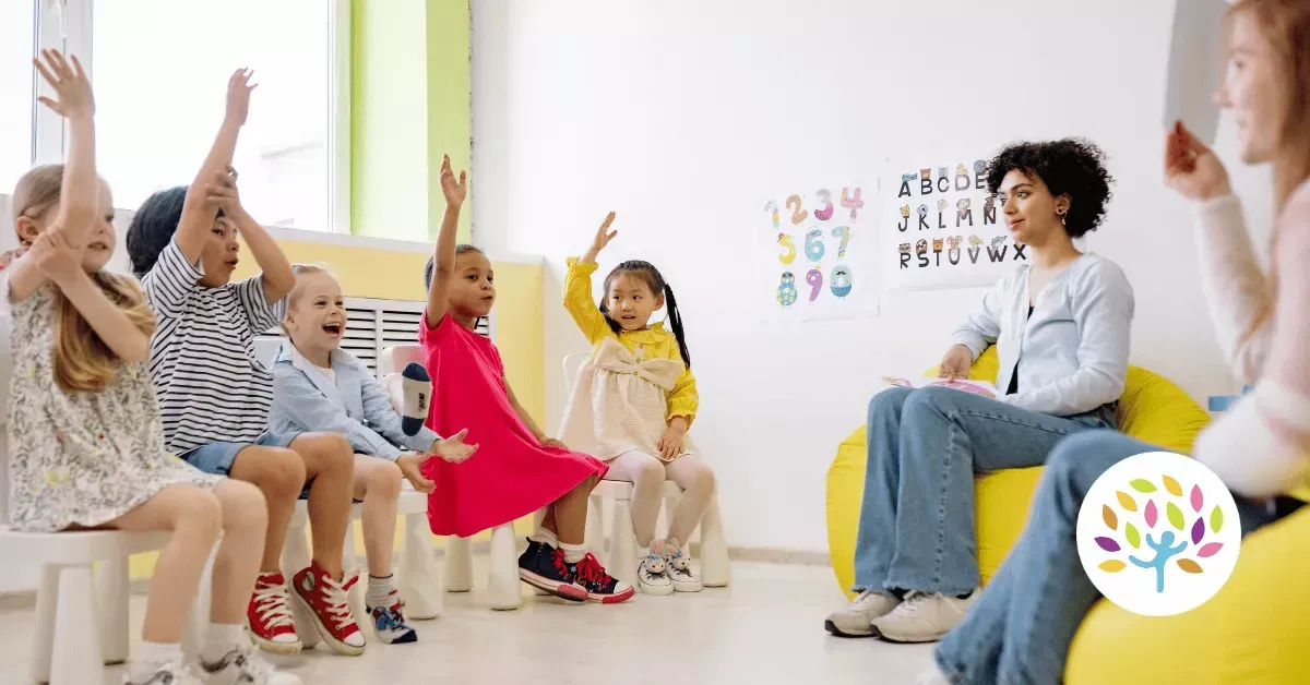 A teacher sits on a beanbag chair in a classroom while students seated in chairs raise their hands to participate.