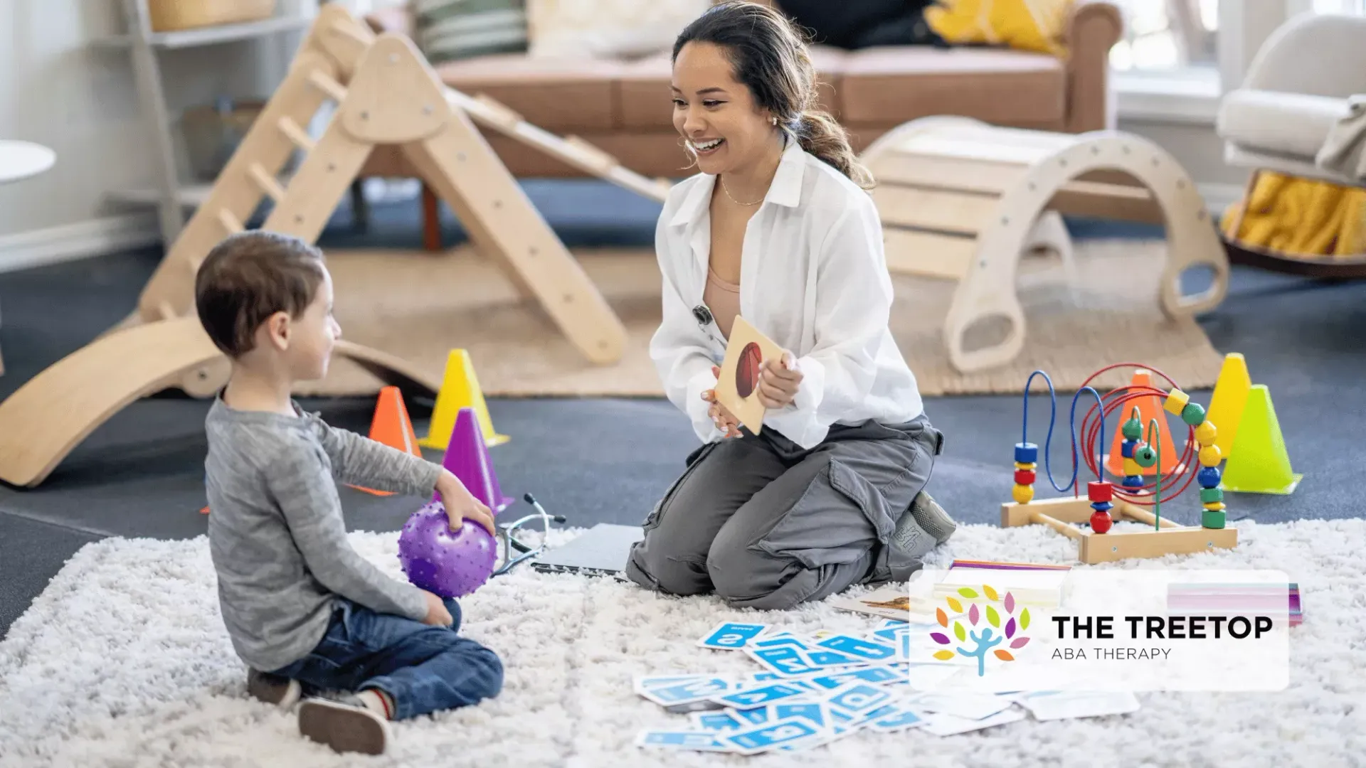 An educator and child sit on a rug in a playroom, engaging in a learning activity with cards and colorful toys.