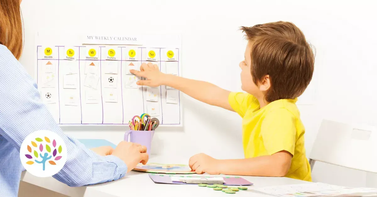 A child and a person work together at a table, using a weekly activity planner chart on the wall.