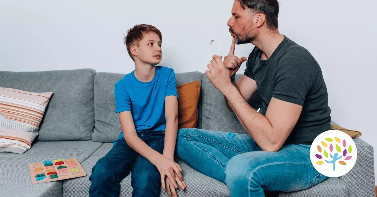 A seated adult gestures toward their mouth while talking to a young person on a couch, with a small puzzle board nearby.