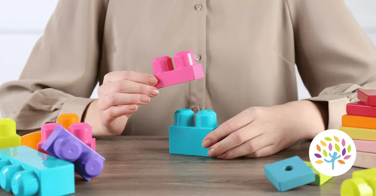 A person’s hands are placing a small pink plastic building block onto a larger blue block on a wooden surface.