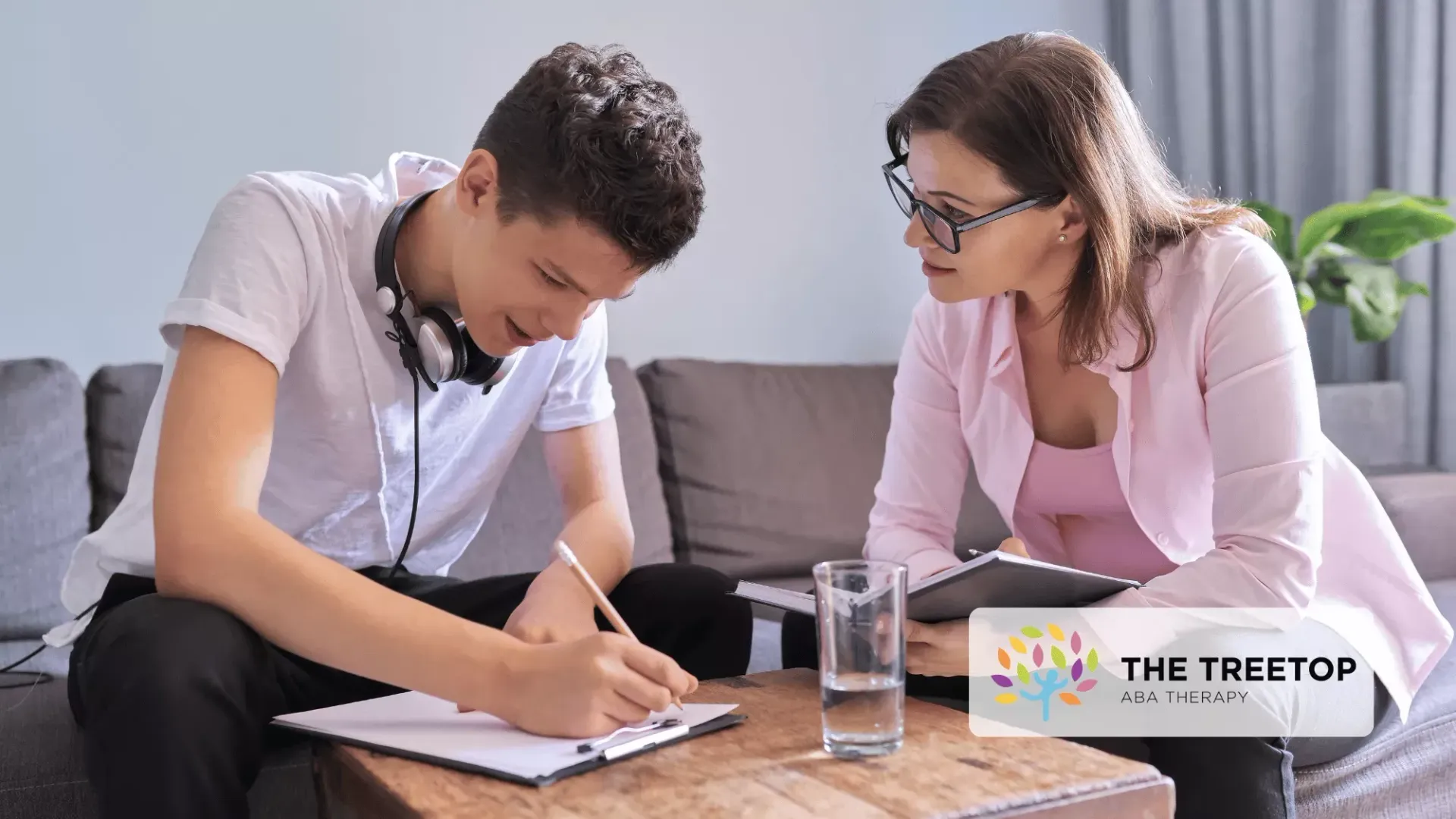 A tutor and student sit on a sofa, looking at a notepad on a coffee table while working together.
