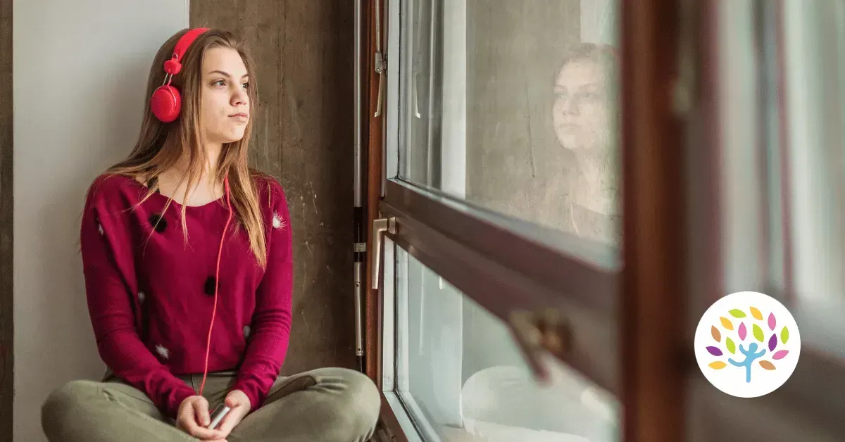 A person in a maroon sweater and headphones sits by a window, gazing outside with a neutral expression.