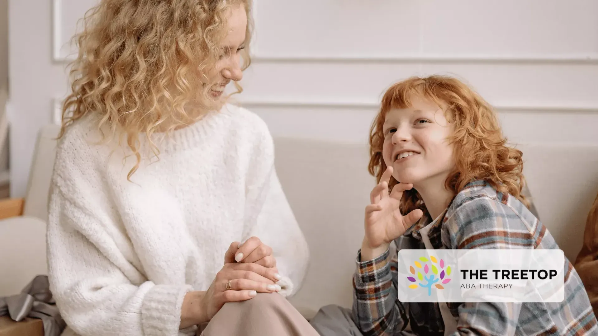 A person with curly hair smiling and talking with a child in a plaid shirt on a couch in a bright room.