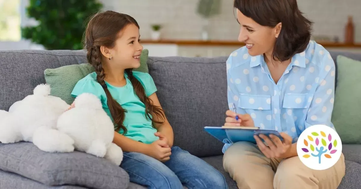 Woman and child sit on a couch, smiling and talking, with a white teddy bear nearby and a blue clipboard in hand