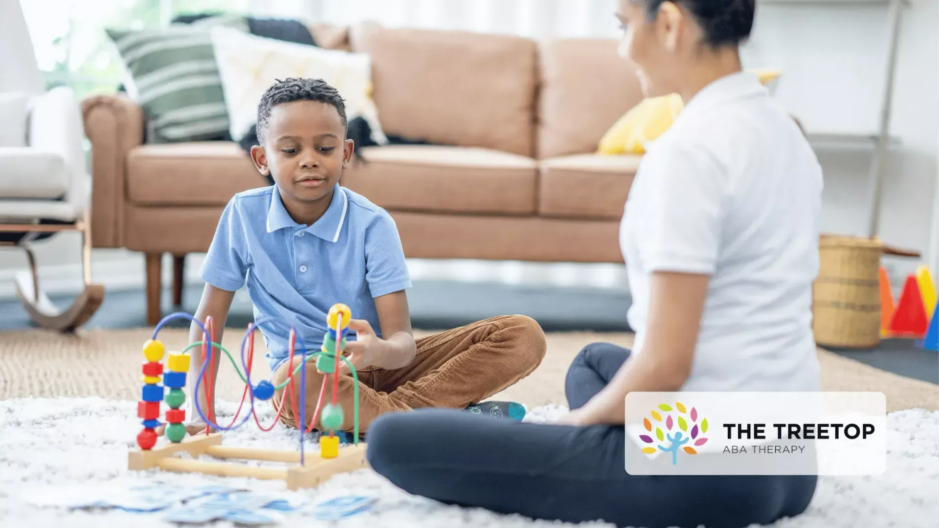 A child sitting on a rug playing with a colorful bead maze toy while a person sits nearby in a bright, modern room.