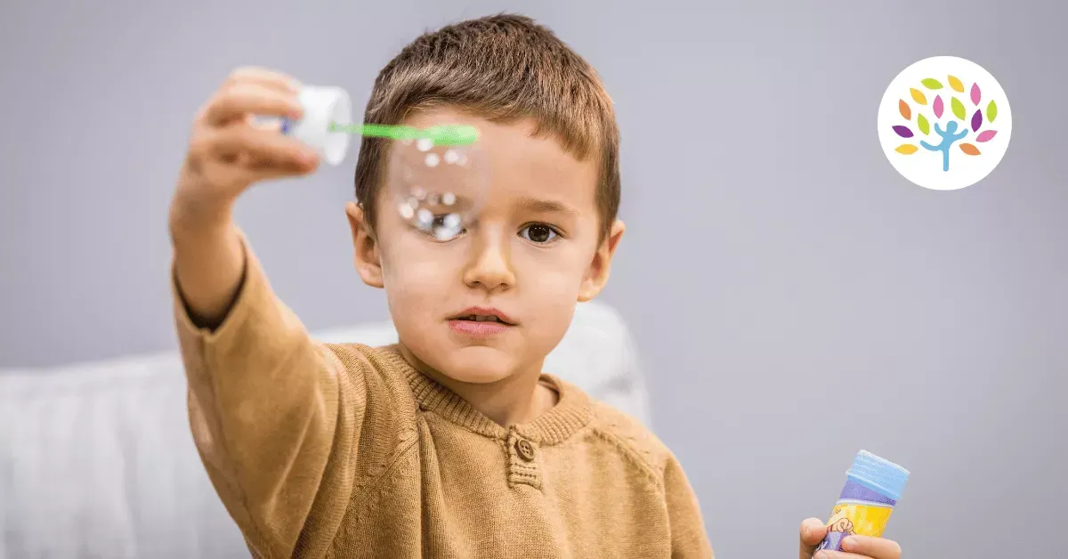 A young child in a tan sweater plays with a bubble wand, holding it near their face while holding a bubble container.