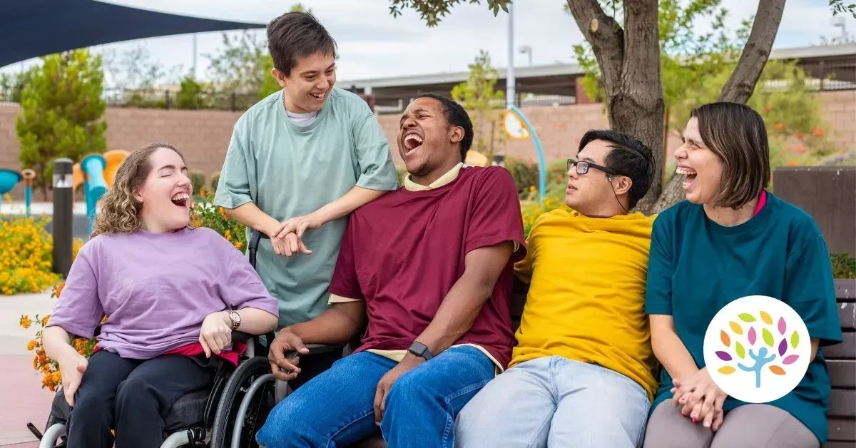 Five diverse people laughing together on a park bench, including one person in a wheelchair and a colorful logo.
