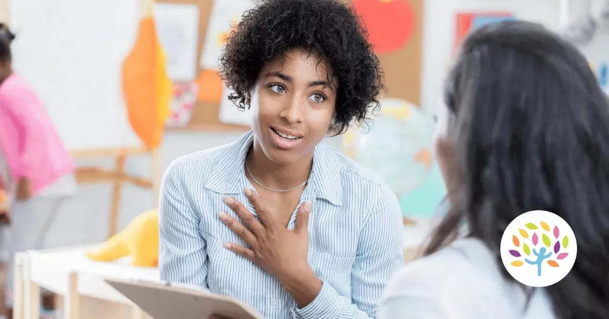 A person with dark, curly hair gestures toward their chest while speaking to another person in a classroom setting.