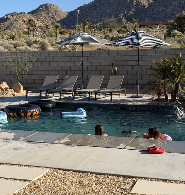 Two people swim in a backyard pool in a desert setting with lounge chairs and umbrellas on the patio.