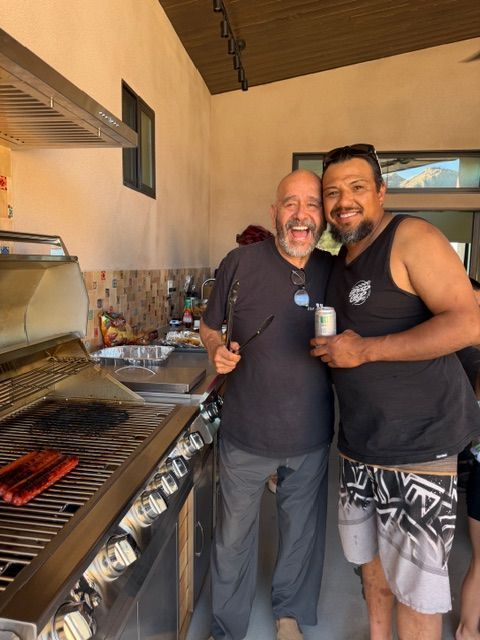 Two men smiling and embracing next to a stainless steel grill cooking meat on an outdoor patio.