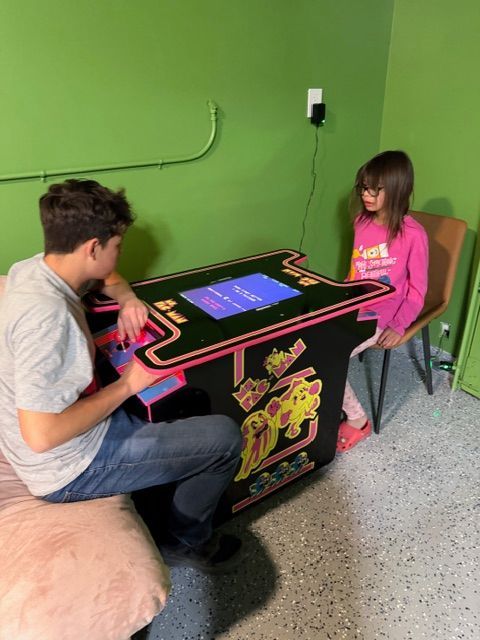 Two people sit opposite each other at a cocktail-style arcade machine in a room with bright green walls.