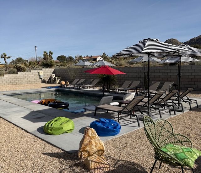 A sunny outdoor swimming pool area with lounge chairs, umbrellas, and colorful pool floats in a desert setting.