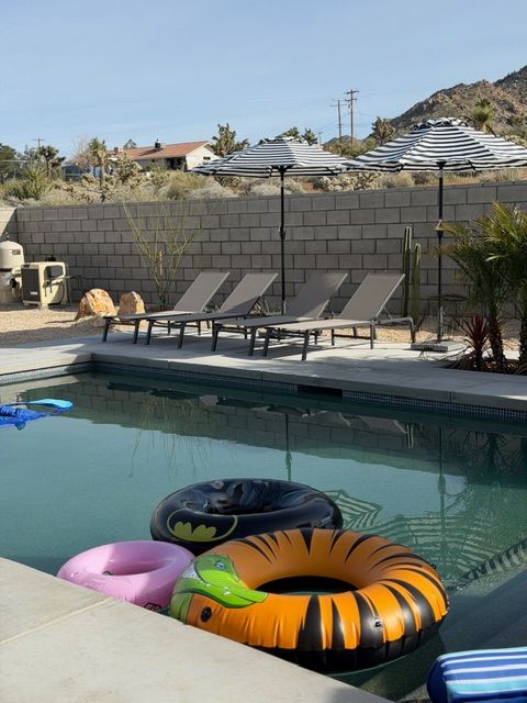 Pool with three floating rings, lounge chairs, and two umbrellas against a block wall in a desert setting.