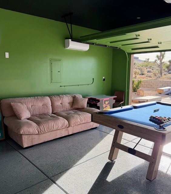 A garage conversion features a light pink sofa, a blue pool table, and green walls, opening onto a desert landscape.