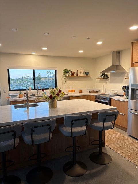 Modern kitchen with a white island, four grey bar stools, wood cabinets, and a window overlooking a desert landscape.