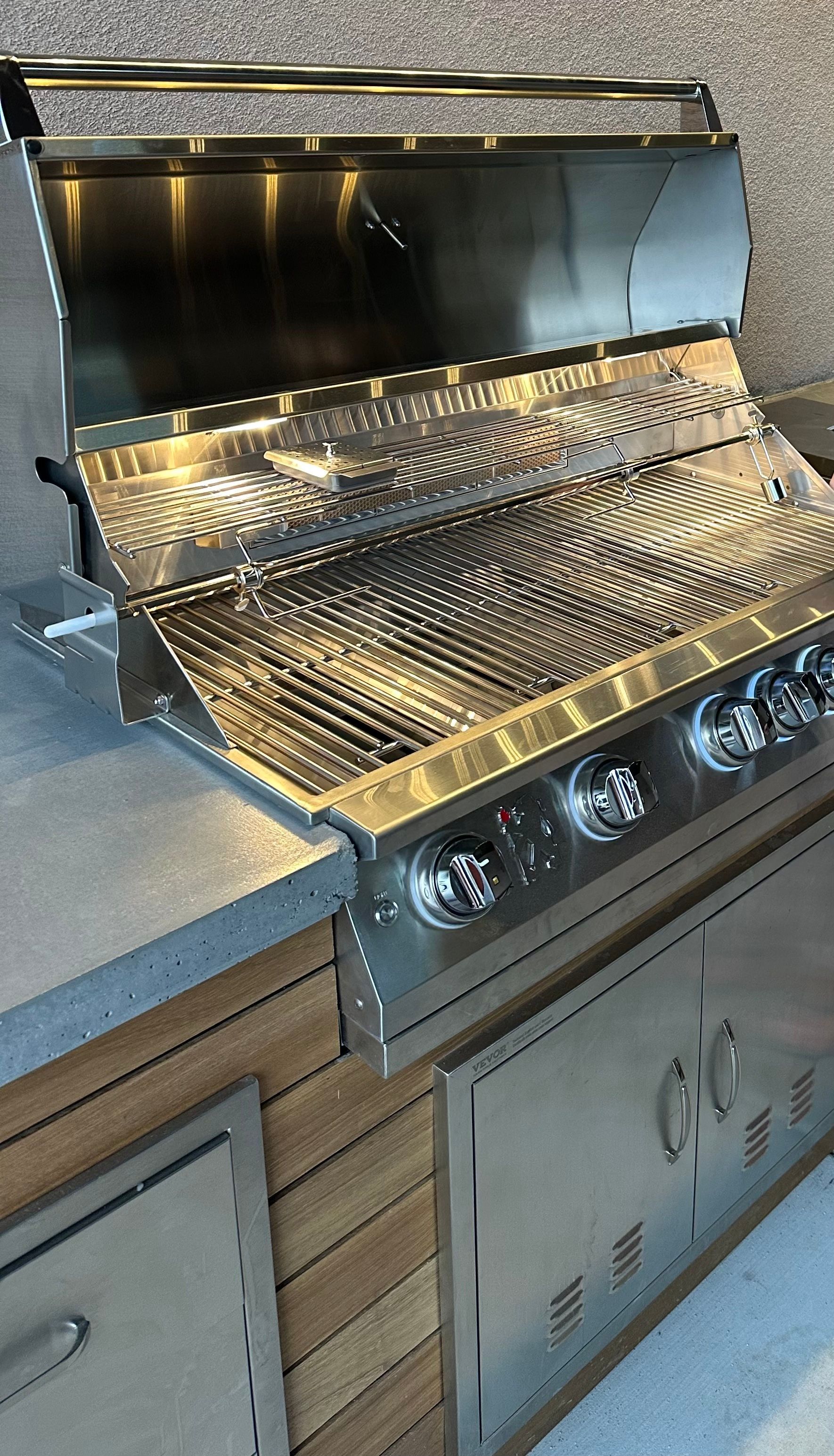 Stainless steel outdoor grill with open lid, set on a countertop with cabinet storage.