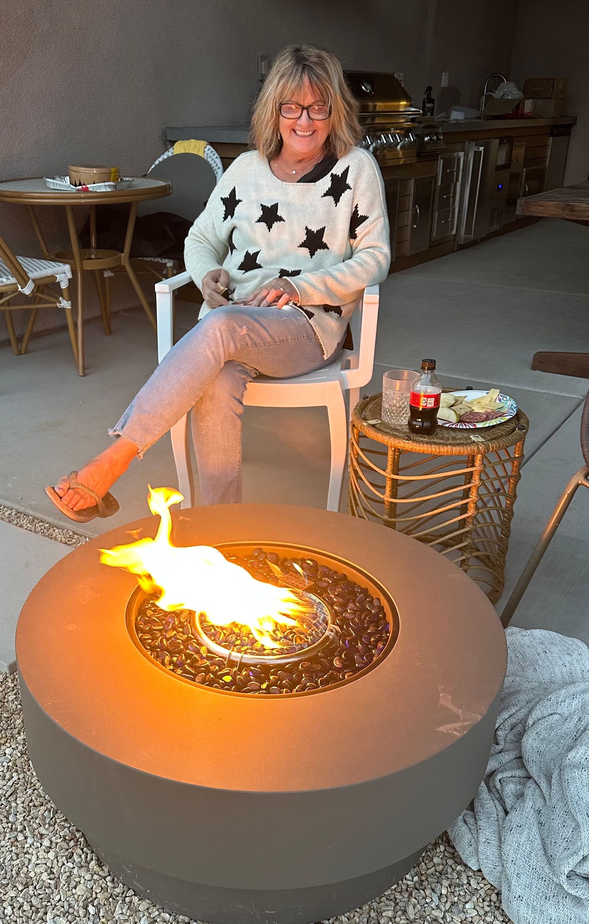 Woman seated near a fire pit, wearing a star sweater and jeans, with a drink on a side table.