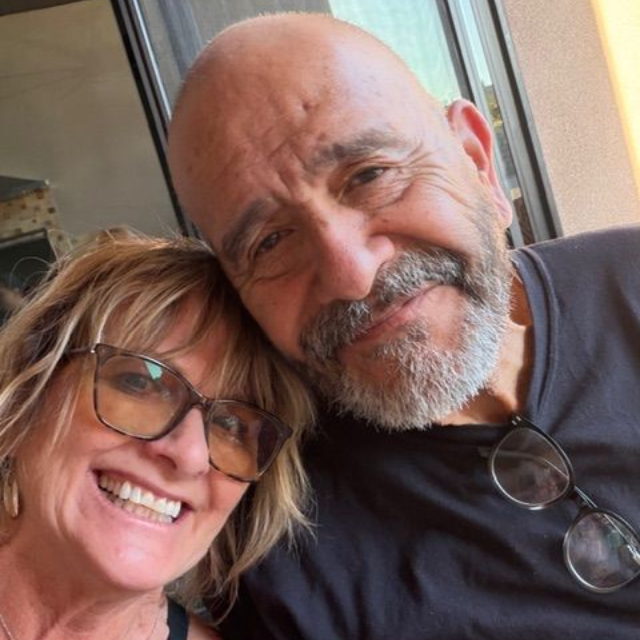 A smiling woman with glasses leaning her head against a man with a gray beard, both looking toward the camera.