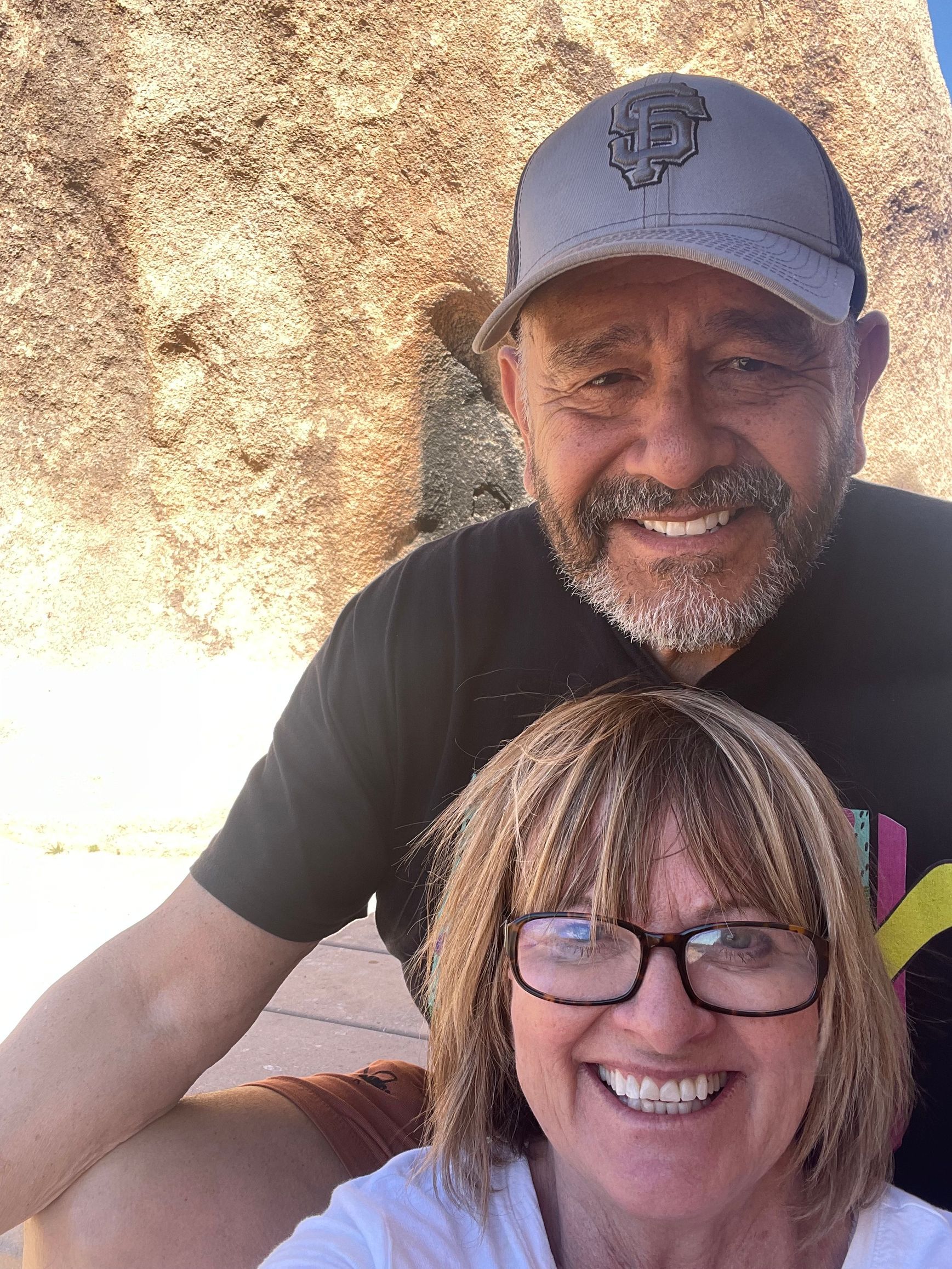 A smiling couple poses for a selfie in front of a light-colored, textured stone wall in bright daylight.