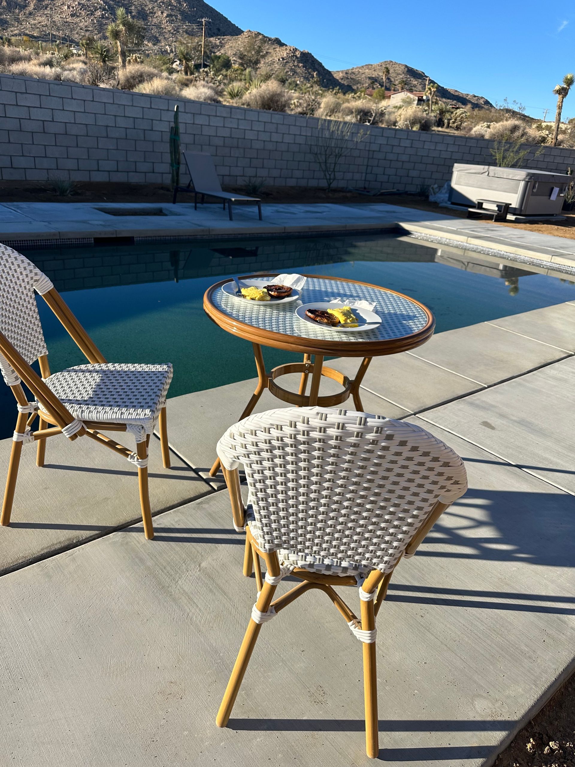 Two white woven chairs, round table with treats, by a pool in a desert setting.