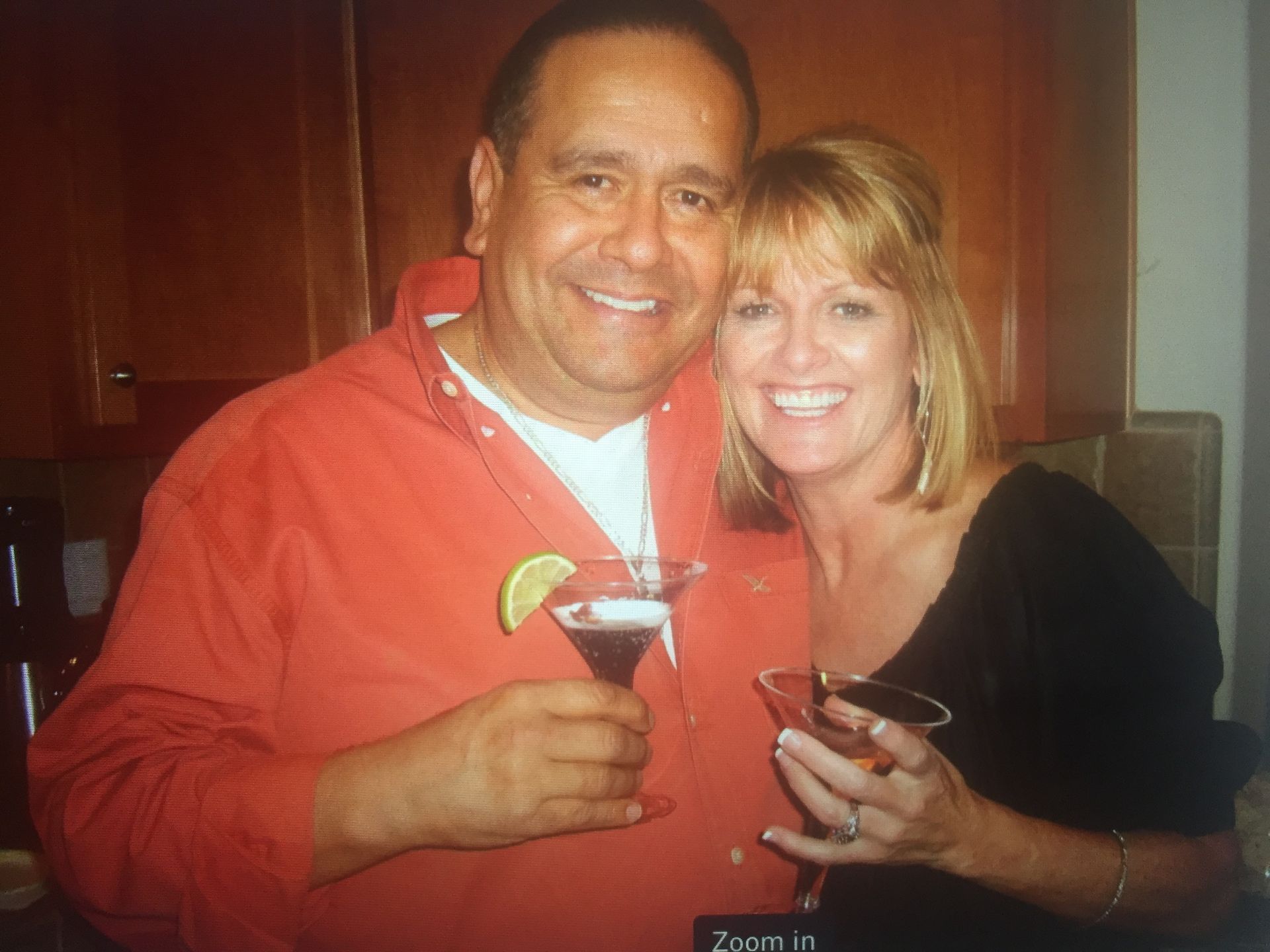 A man in an orange shirt and a woman in a black shirt smiling while holding martini glasses in an indoor kitchen setting.
