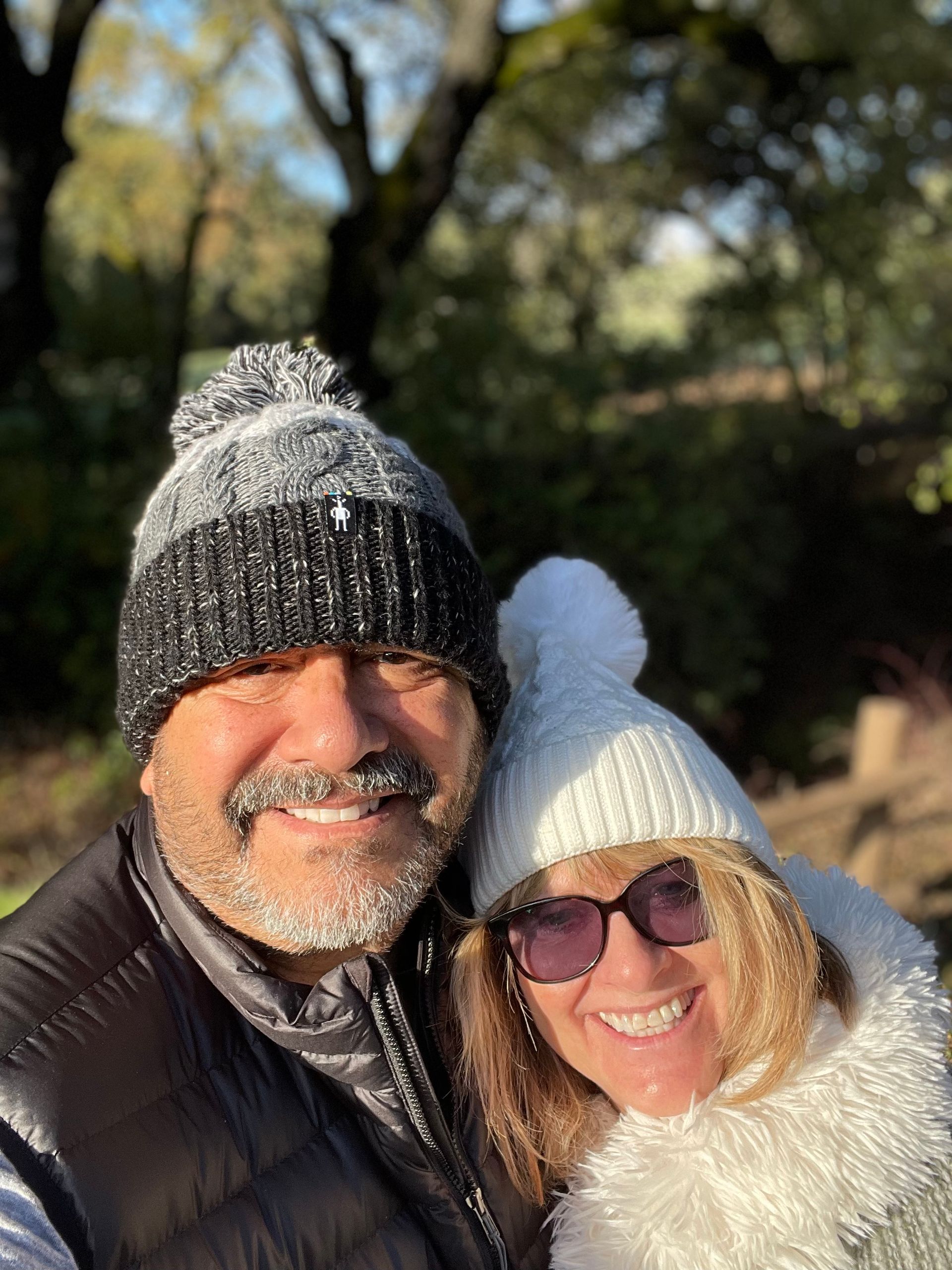 A smiling couple in winter hats and scarves poses for a selfie outdoors with trees in the background.