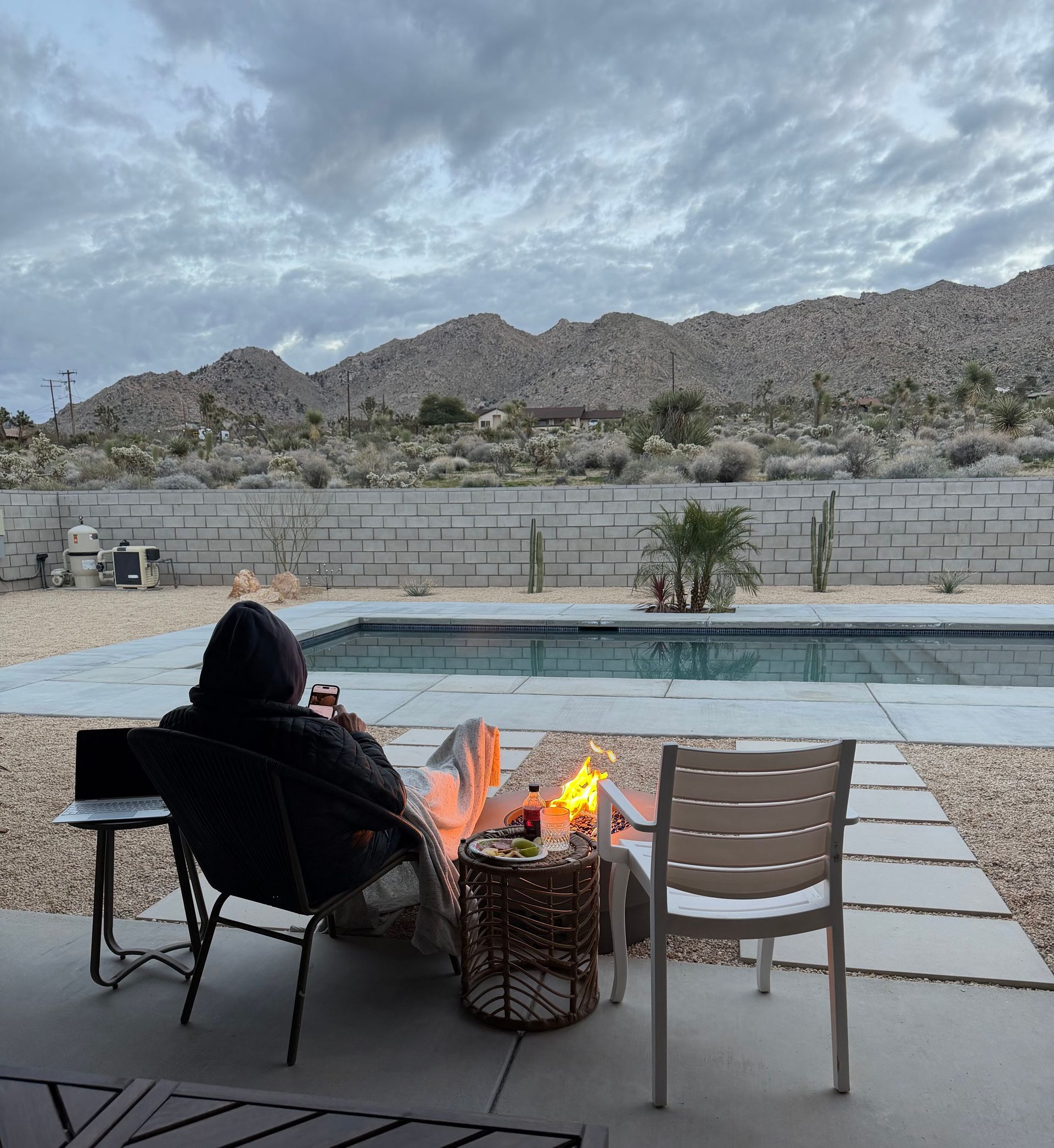 Person seated by a fire pit, overlooking a pool and mountains. Overcast sky.