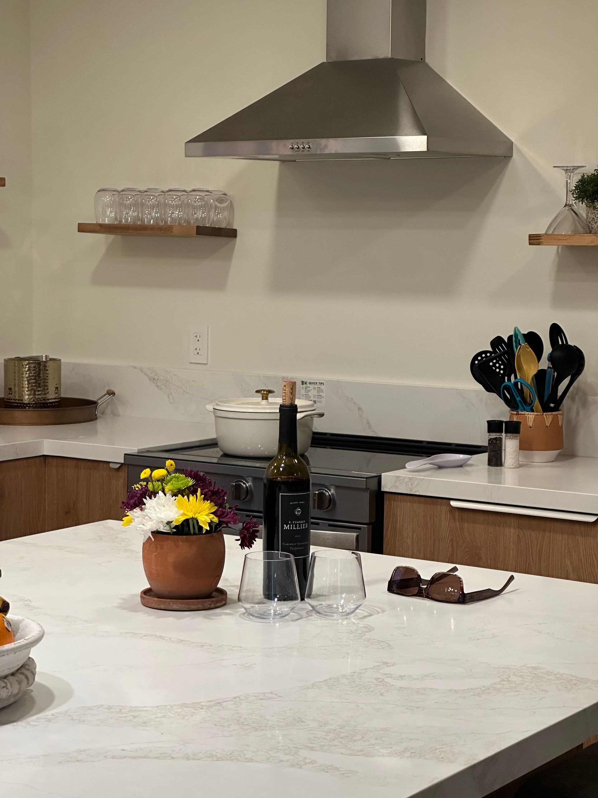 Kitchen counter with flowers, wine bottle, and sunglasses. Stainless steel range hood above stovetop.