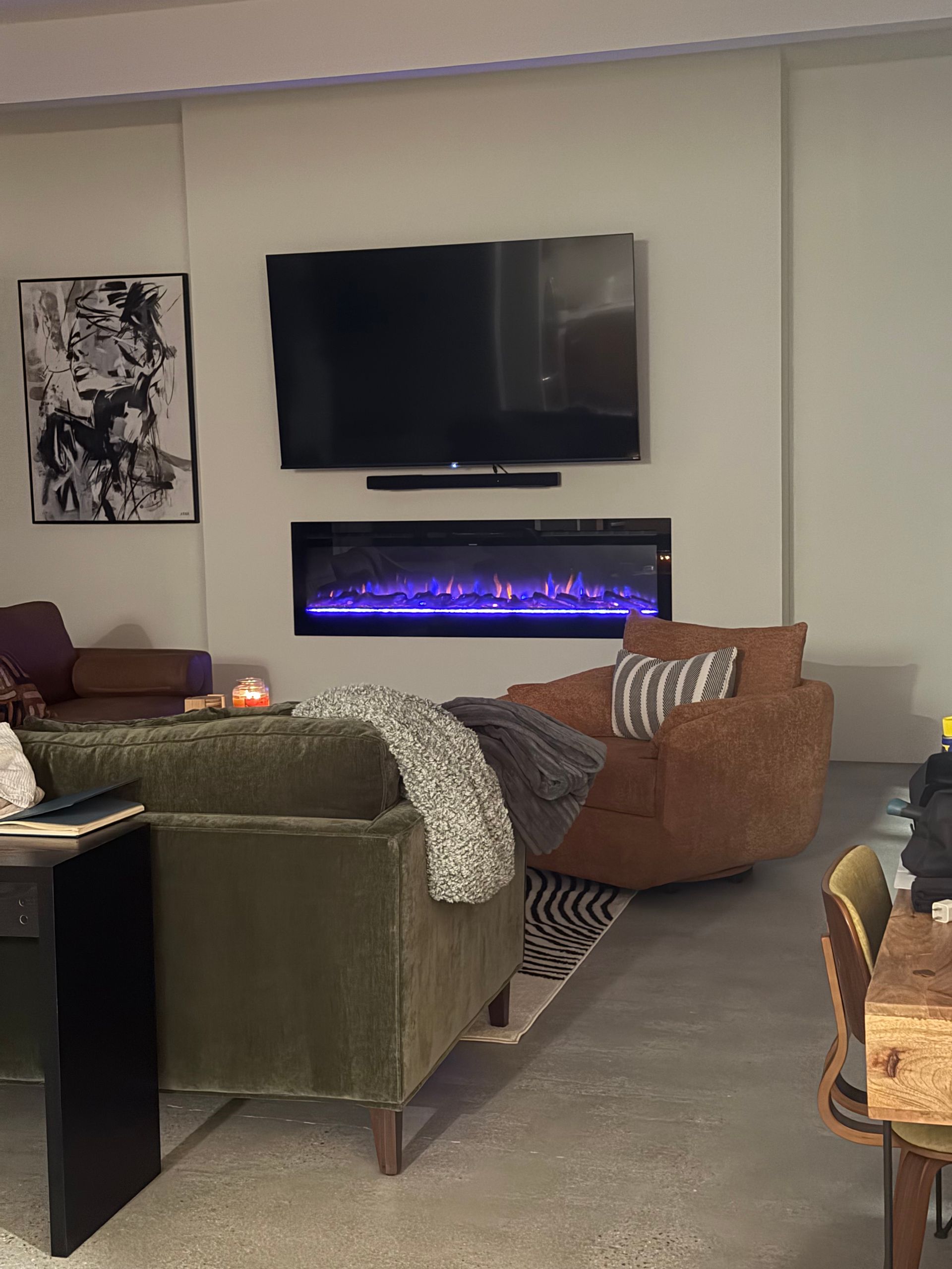 Living room with TV above a fireplace, green couch, brown chair, art, and rug.