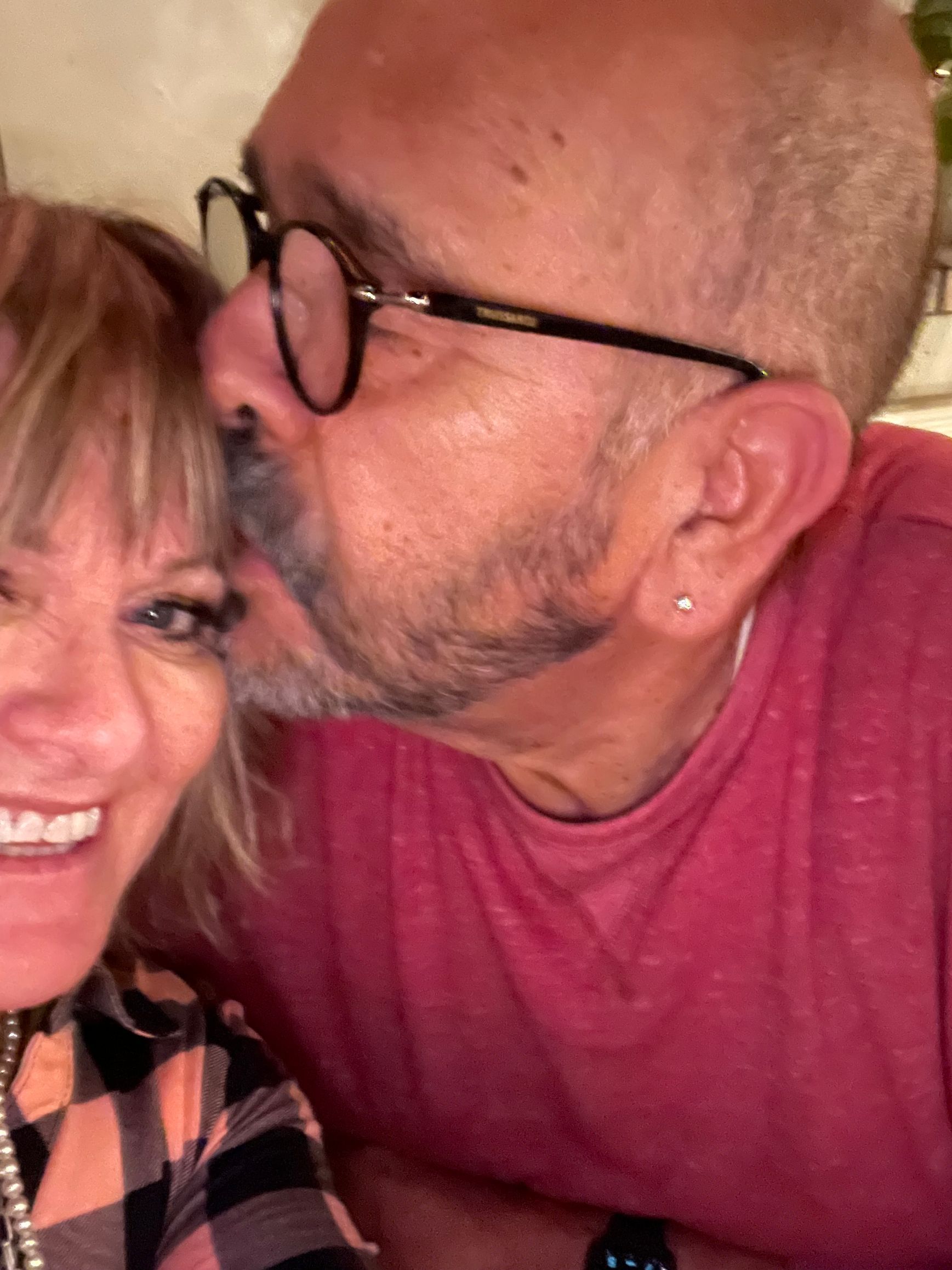 A person in a red shirt kisses the forehead of a smiling person wearing a plaid shirt.