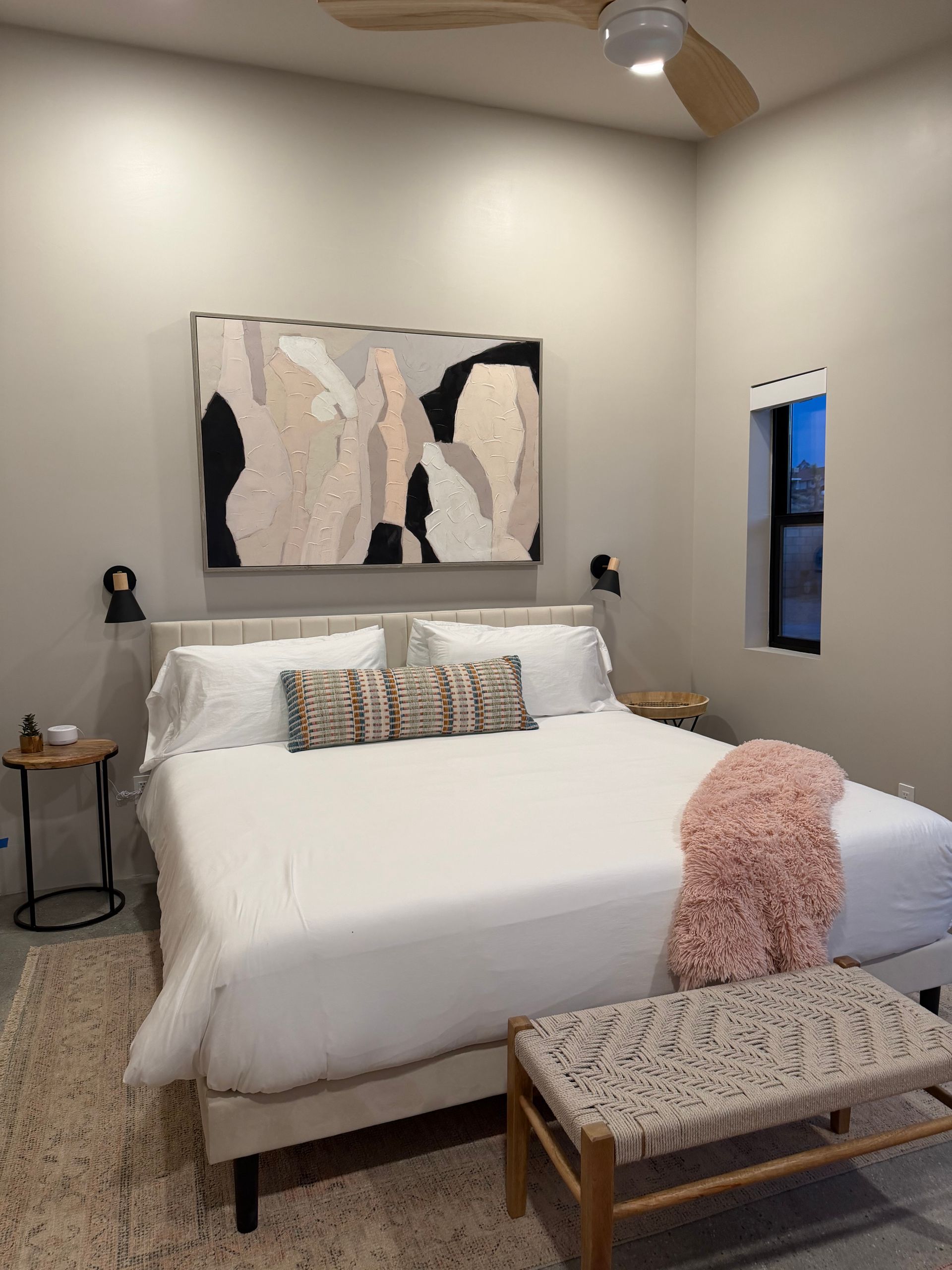 Bedroom with large abstract art, bed, bench, and side table. Pale pink, white, and black colors.