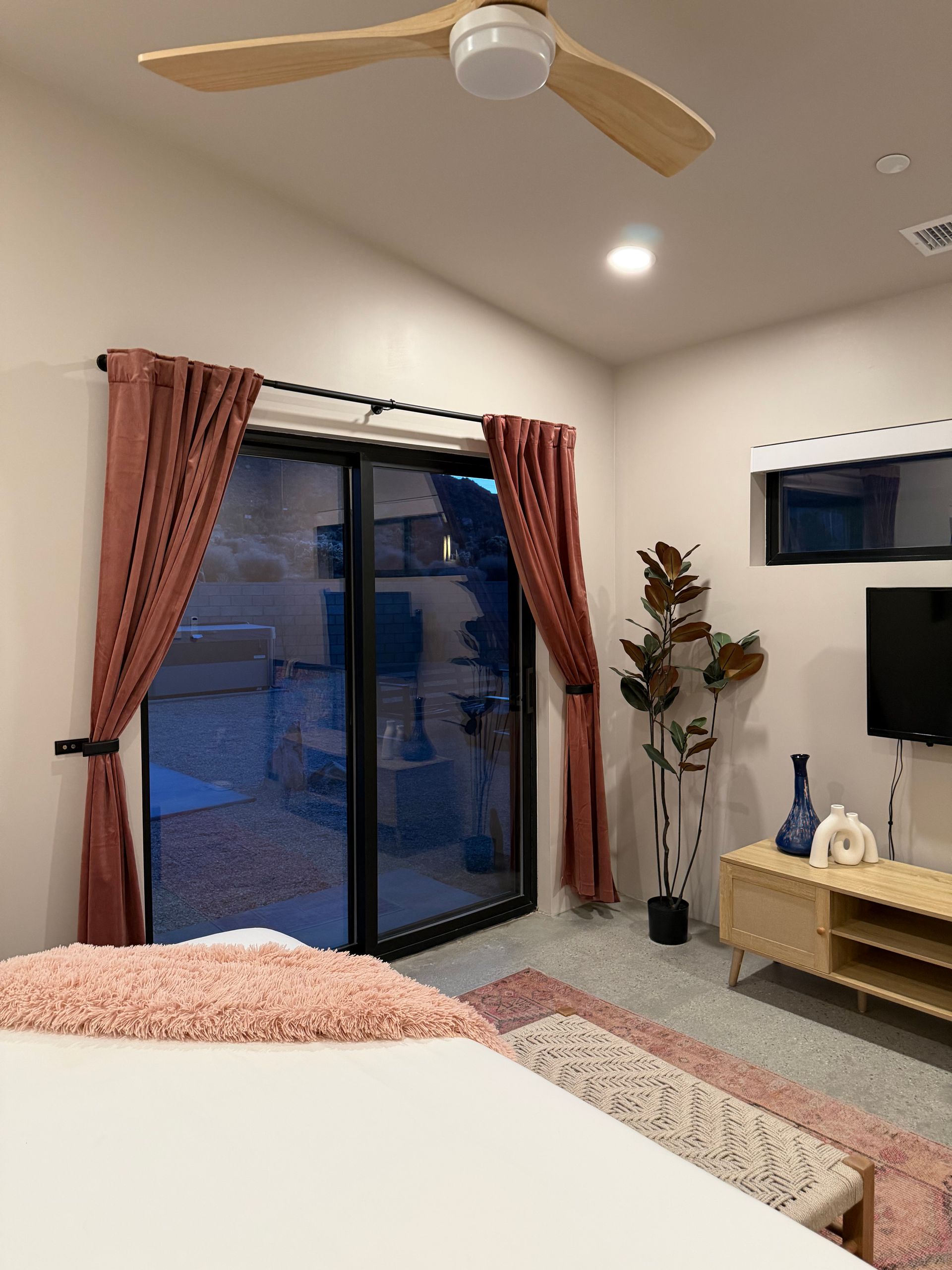 Bedroom with sliding glass door, mauve curtains, ceiling fan, TV, and light wood furniture.