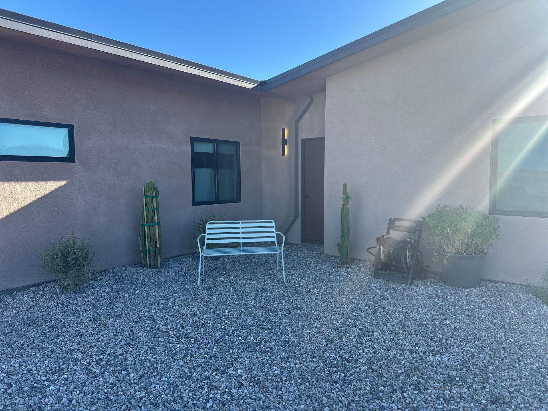 Exterior view of a house with a gravel yard, bench, and plants. Light brown stucco walls, dark windows.