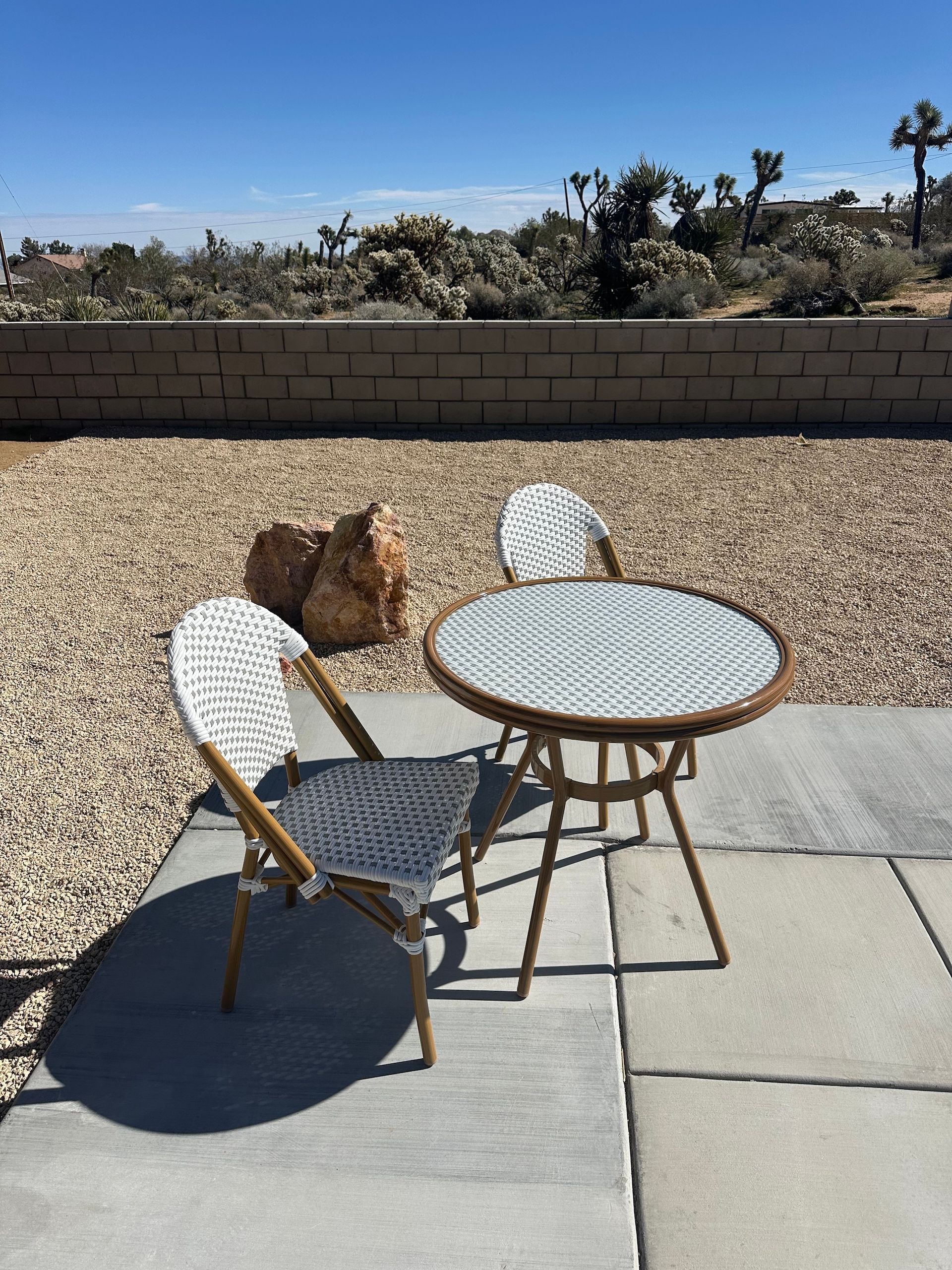 Patio set with white woven chairs and round table, set on concrete pavers, surrounded by gravel and desert landscape.