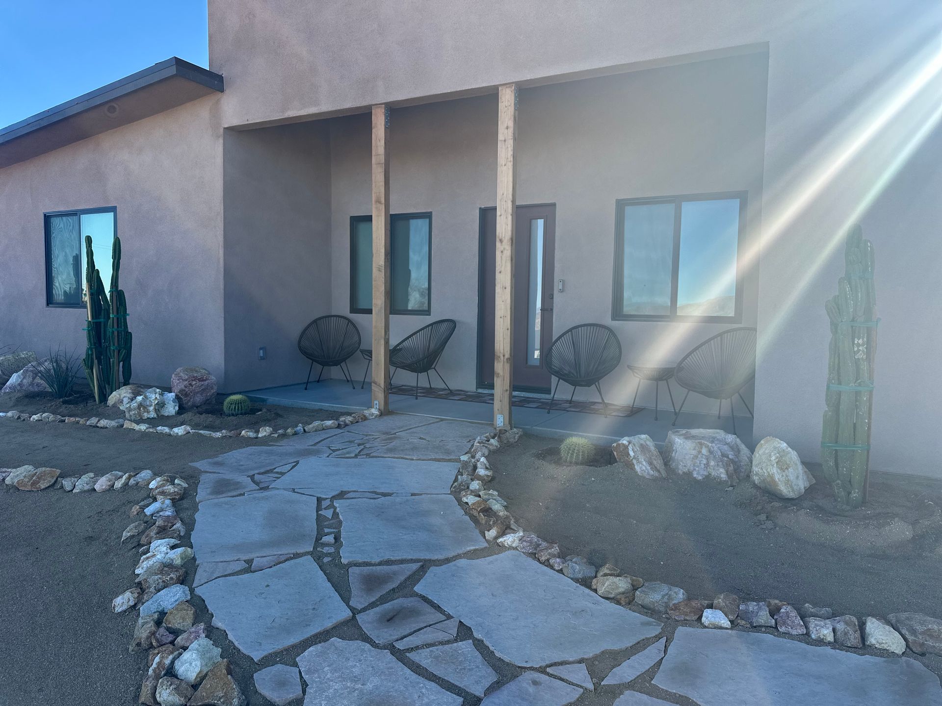 Stone path leads to a tan stucco house with porch, two chairs, and cacti.