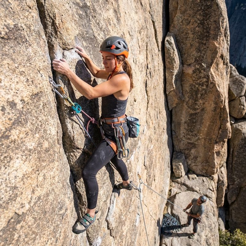 A rock climber ascends a granite cliff face, secured by gear, with a belayer standing on a ledge below.